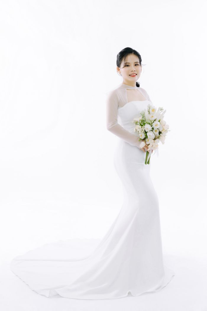 About Studio portrait of a bride in a white wedding dress holding a bouquet, against a plain background.