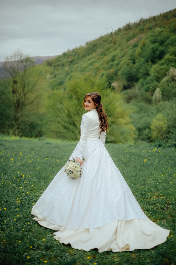 About A bride poses elegantly in a white wedding dress with a bouquet against a lush green hillside.