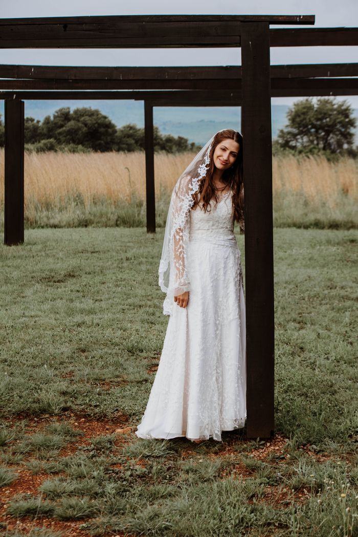 About A beautiful bride in a lace wedding dress poses outdoors with wooden pergola.