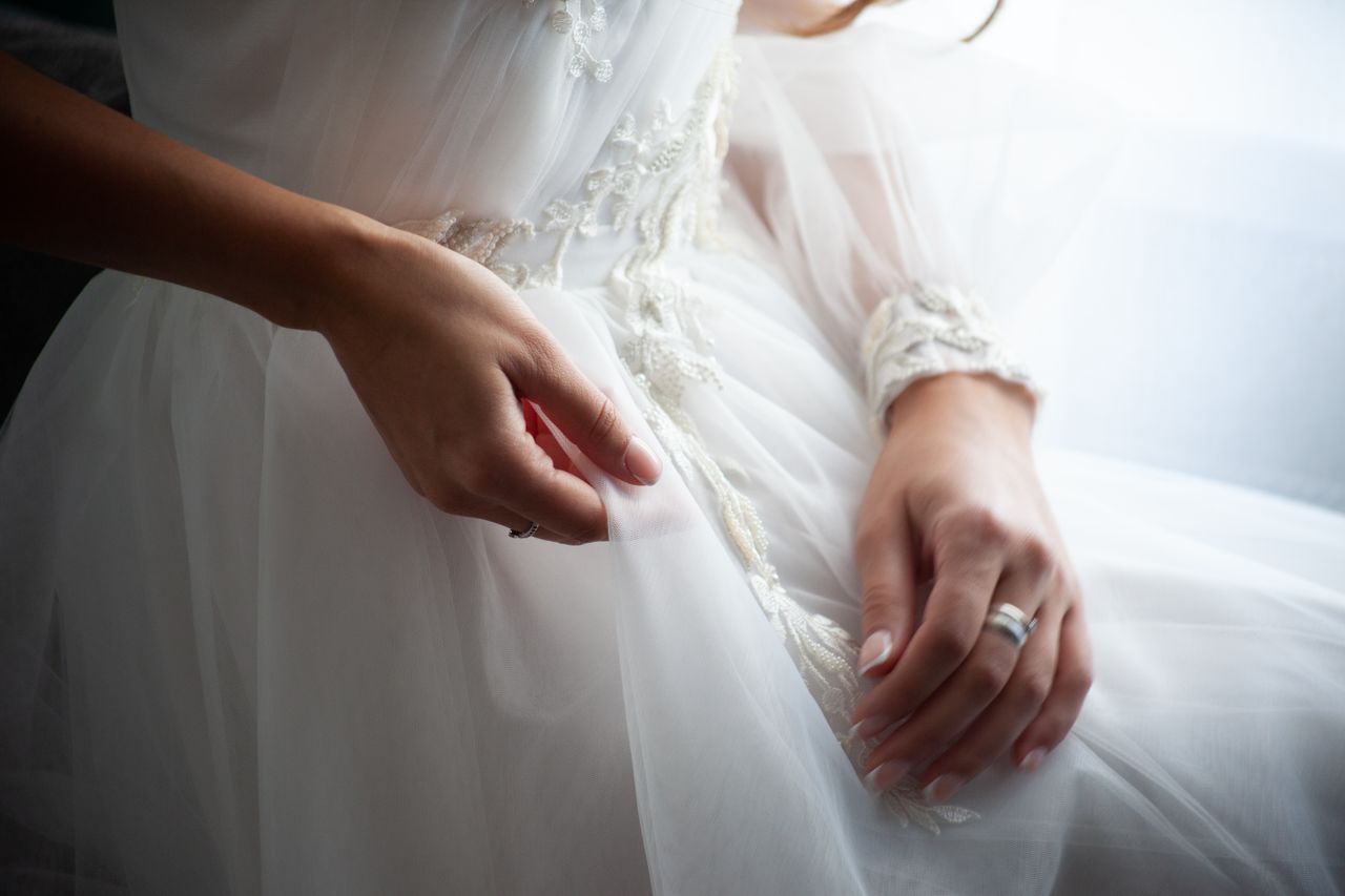 Contact A bride's hands gracefully resting on her elegant wedding dress, showcasing delicate details.